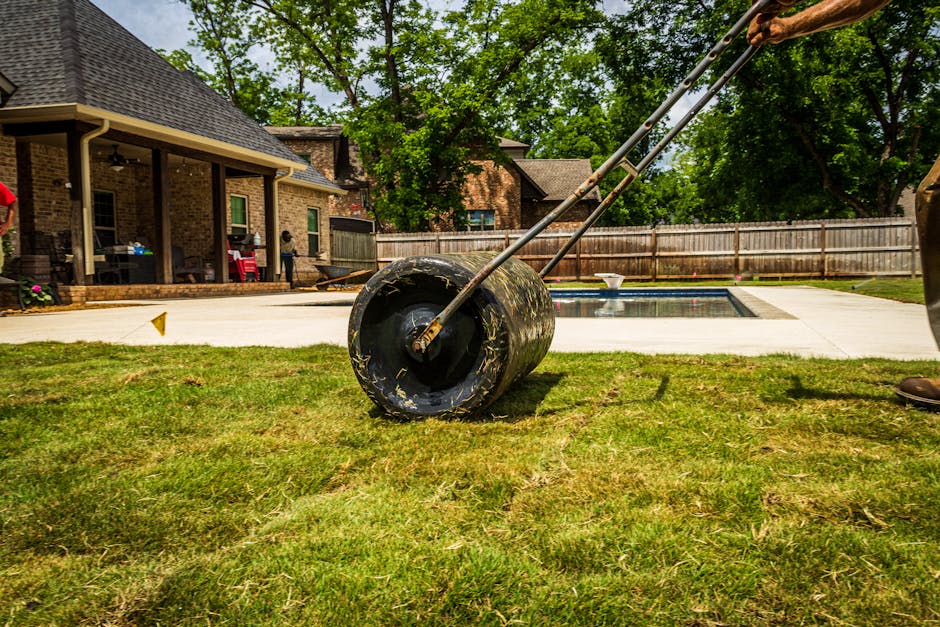 A person wearing a plaid shirt, jeans, and boots is kneeling on a garden lawn, preparing the soil for new turf or planting. They are rolling out a thick layer of sod, exposing the soil beneath, which 