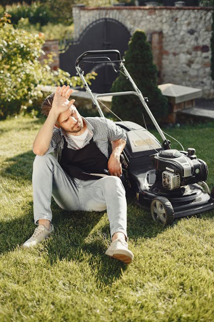 A young man seated on a well-maintained lawn, appearing to rest after gardening work, with a petrol-powered lawn mower placed beside him. The garden features neatly trimmed grass, a stone wall with an